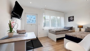 Ein modernes Zimmer mit einem Bett, einem Schreibtisch, einem Stuhl, einem an der Wand befestigten Fernseher und einem Fenster mit Vorhängen.