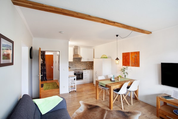 A modern living room with a sofa, a dining table with chairs, a kitchen in the background, and a flat-screen TV mounted on the wall.