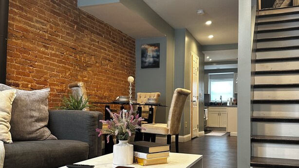 A modern living room with a brick wall, a sofa, a coffee table, and a staircase leading to another level. - Cozy 2BR Retreat with Free Parking & Prime Location (Baltimore)