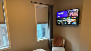 A hotel room with a bed, a chair, a TV displaying a gaming interface, and windows with blinds. - Cozy 2BR Retreat with Free Parking & Prime Location (Baltimore)