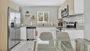 A modern kitchen with a stainless steel refrigerator, built-in oven, and microwave. There is a window with blinds, a sink, and a countertop with a plant.