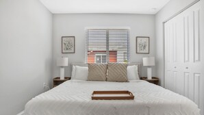 A neatly arranged bedroom with a large bed, two bedside tables with lamps, a window with blinds, and a door.
