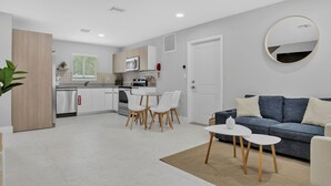 A modern kitchen with white cabinets, a dining area with white chairs, and a grey sofa with white cushions.