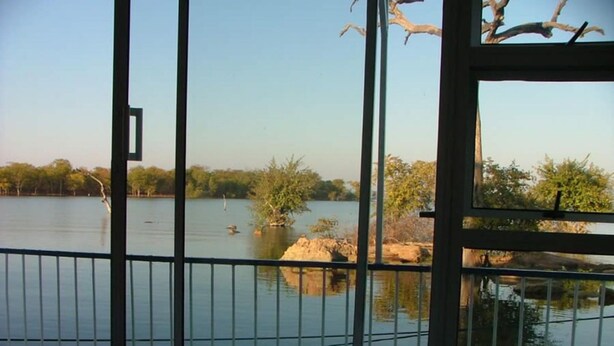 A room with a view of a calm lake, a metal railing, and a window with a view of trees and a clear sky. - Houseboat With Aircon and Splash Pool - 2128 (Kariba)