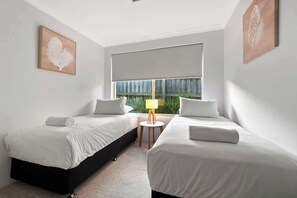 A hotel room with two beds, a small table with a lamp, and a window with a view of greenery.