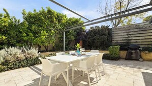 Un patio con mesa y sillas blancas, rodeado de follaje y una parrilla.