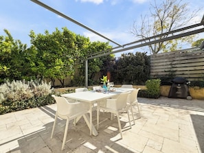 Une terrasse avec une table et des chaises blanches, entourée de verdure et agrémentée d’un barbecue.