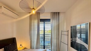Una habitación de hotel moderna con una cama grande, un ventilador de techo, una ventana con cortinas, un radiador fijado a la pared y un cuadro enmarcado.