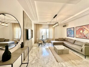 A modern living room with a marble floor, a large round mirror, a sofa, a coffee table, and artwork on the walls. - Elyssa Superbe Appartement aux Jardins de Carthage (Tunis)