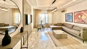 A modern living room with a sectional sofa, a round coffee table, a dining area with a glass table, and artwork on the walls. - Elyssa Superbe Appartement aux Jardins de Carthage (Tunis)