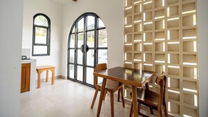A dining area with a wooden table and chairs, a small wooden stool, and a large window with a view outside. - Kanchana Cottages (Pecatu)