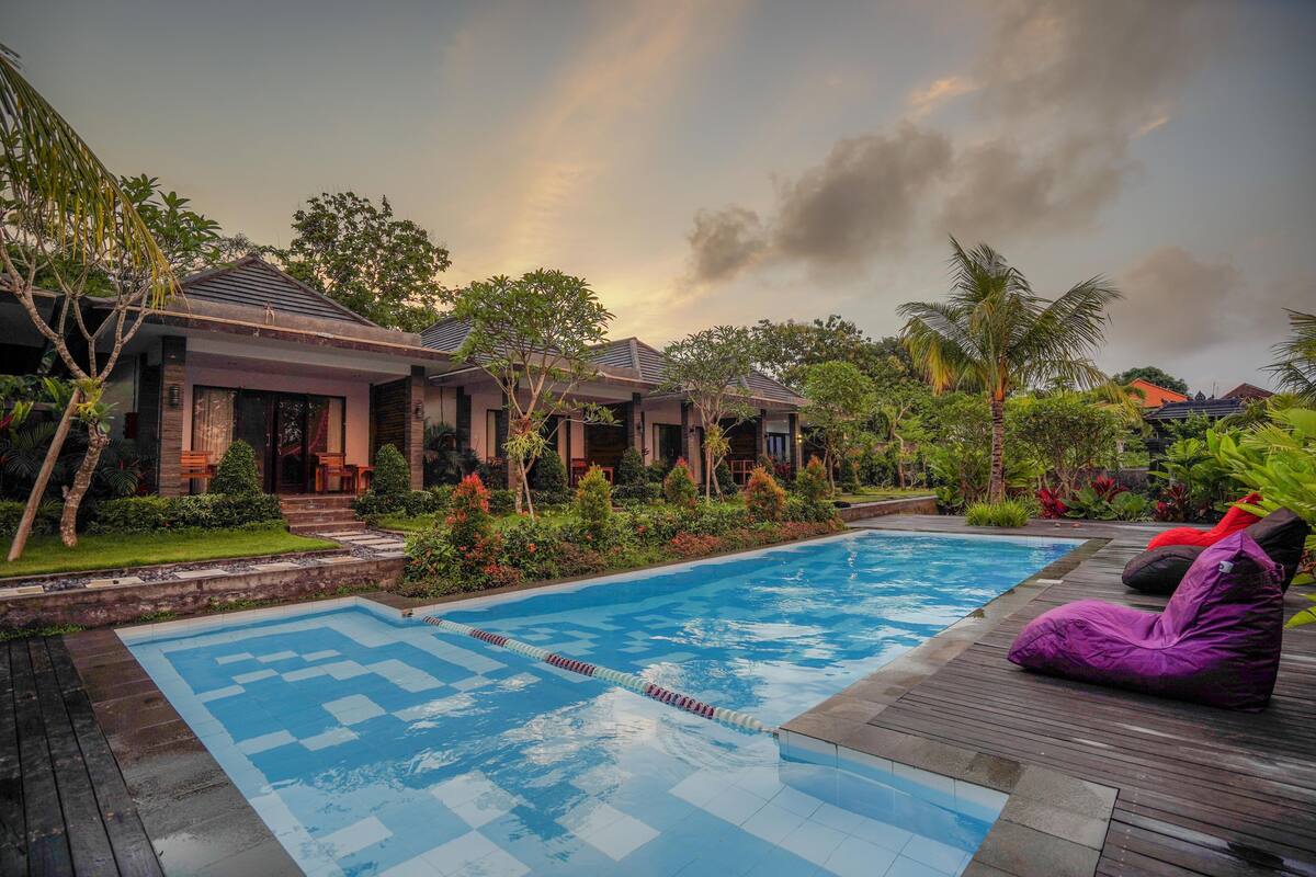 A swimming pool with a wooden deck and lounge chairs, surrounded by lush greenery and a modern house.