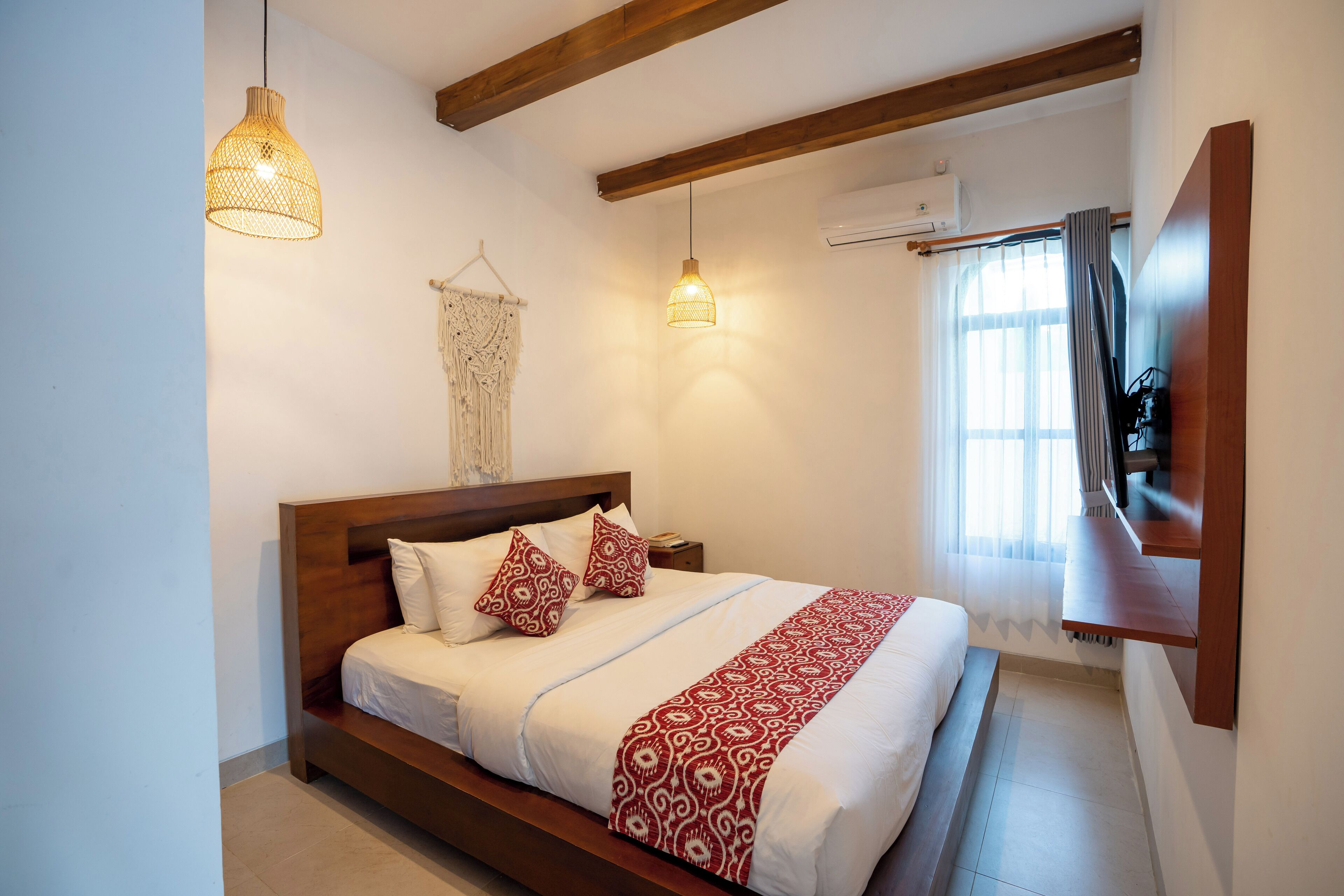 A bedroom with a wooden bed, white bedding, a red and white patterned throw, two hanging pendant lights, a wall-mounted TV, and a window with curtains.