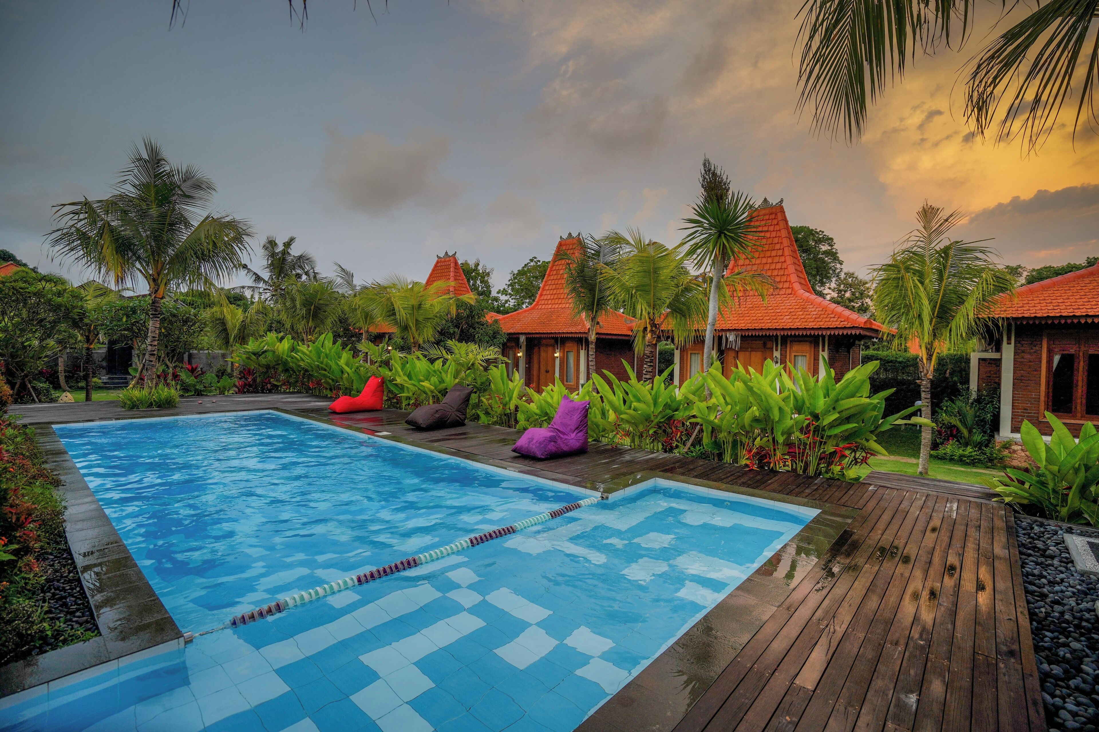 A swimming pool with a wooden deck, surrounded by lush greenery and traditional-style buildings with red roofs.