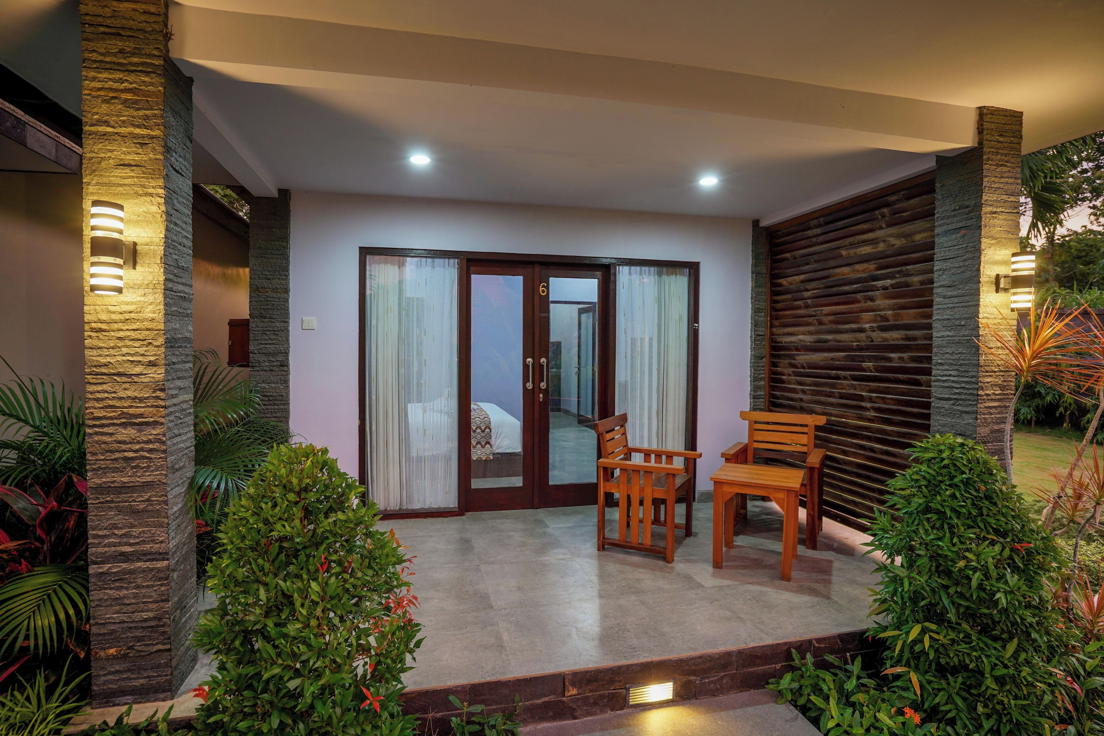 A covered outdoor area with a sliding glass door leading to a bedroom, wooden furniture, and stone pillars.