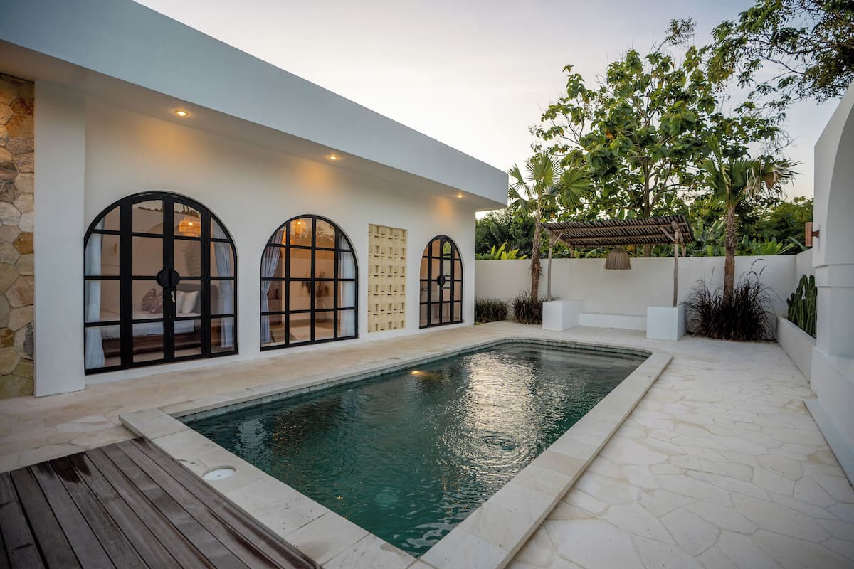 A modern outdoor pool area with a stone deck, a wooden deck, and a white wall with large arched windows.