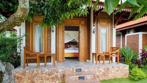 A wooden porch with a bed and two chairs, a stone wall, and a small wooden platform. - Kanchana Cottages (Pecatu)