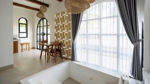 A room with a large window, a wooden dining table with chairs, a white sofa, and a ceiling fan. - Kanchana Cottages (Pecatu)