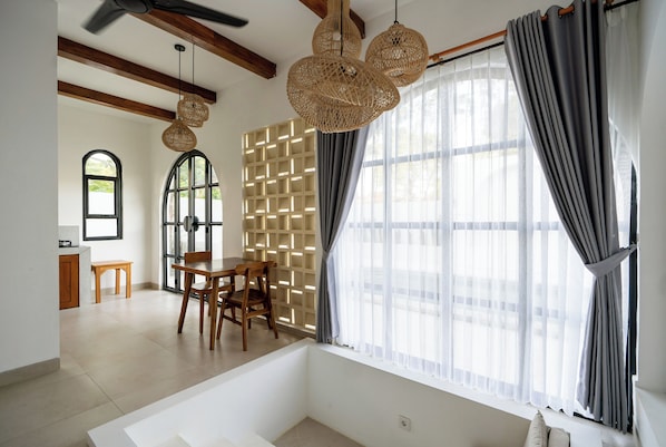 Quarto com uma janela grande, uma mesa de jantar de madeira com cadeiras, um sofá branco e um ventilador de teto.