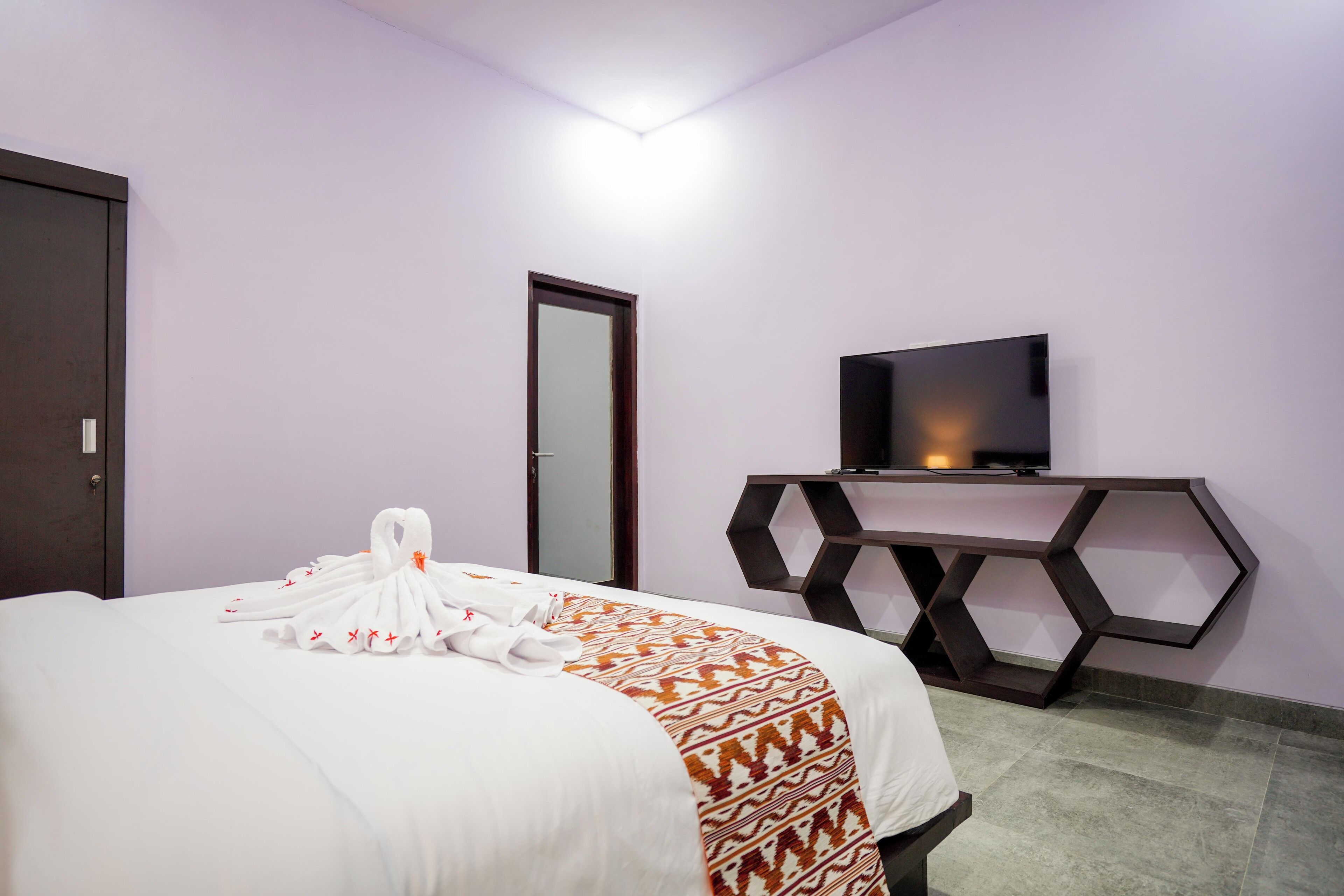 Una habitación de hotel con una cama, un televisor sobre un atril hexagonal y una puerta.