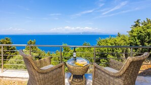 A balcony with wicker furniture, a bottle, and a book, overlooking a coastal landscape. - Villa Ariadne in Apergatika (Paxos)