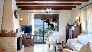 A living room with a fireplace, a TV, and a view of the sea through large windows. - Villa Ariadne in Apergatika (Paxos)