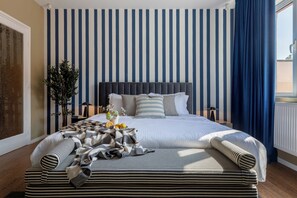A neatly arranged bedroom with a striped wall, a bed with multiple pillows, a bedside table with a lamp, and a window with curtains.