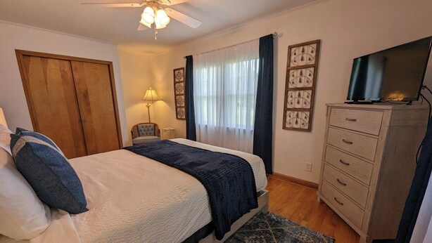 Quarto com cama, cômoda, televisão, ventilador de teto e janela com cortinas.