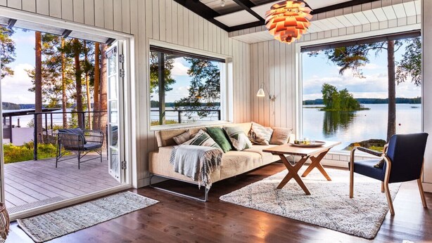 Una sala de estar moderna con sofá, mesa de centro y sillas, ventanas amplias con vista a un lago, y una terraza de madera en el exterior.
