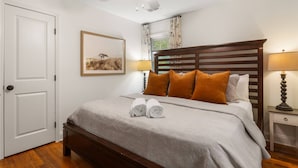 Un lit bien fait, avec une tête de lit en bois, un tableau encadré au mur, un ventilateur de plafond et une table de chevet avec une lampe.