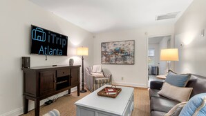 Un salon doté d’un téléviseur à écran plat diffusant l’émission « iTrip Atlanta », d’un meuble de rangement en bois foncé, de fauteuils à rayures, d’une table basse blanche sur laquelle repose un livre, et de deux lampadaires.