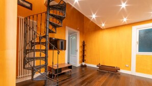 Una habitación con una escalera espiral de madera, un televisor de pantalla plana montado en la pared, un banco de madera y una puerta que da a otra habitación.