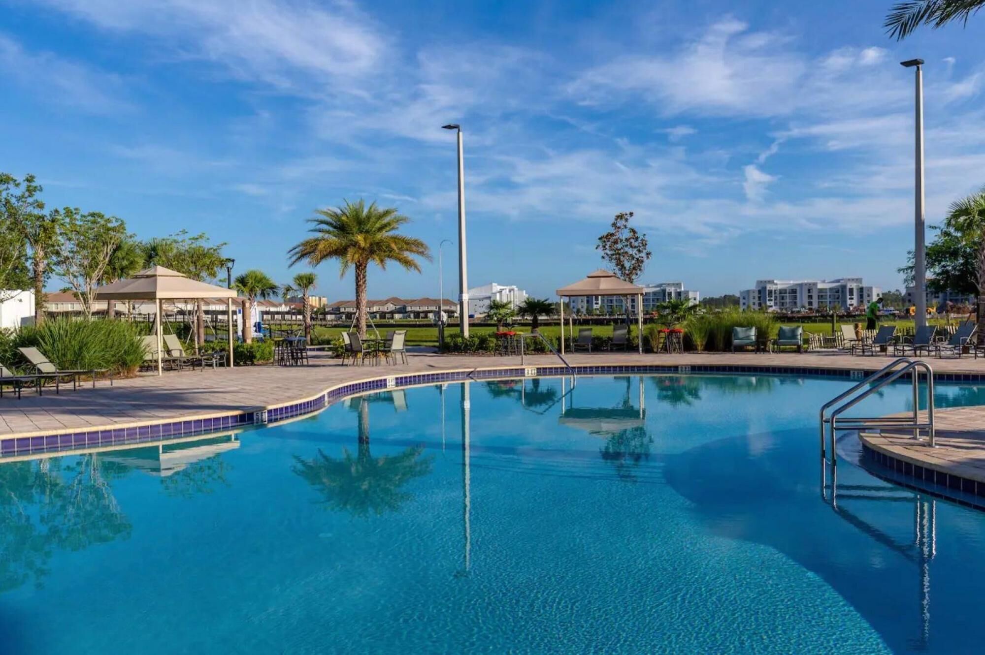 A clear blue pool with lounge chairs and umbrellas, surrounded by palm trees and buildings in the background.