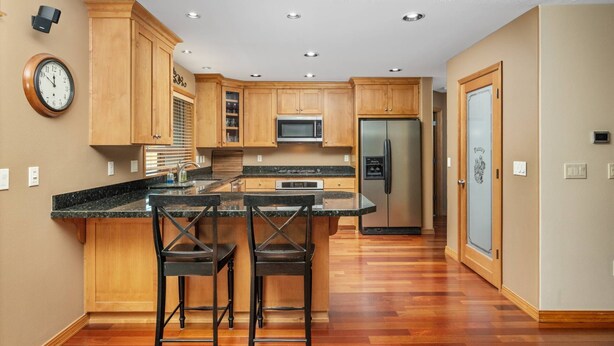 Una cocina con armarios de madera, un refrigerador de acero inoxidable, un microondas, un reloj y una zona de bar con taburetes.