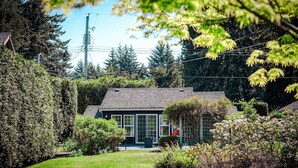 A house with a sloped roof, surrounded by a well-maintained garden and dense trees. - Sit Back & Watch Waves at Rathtrevor Cottage (Parksville)