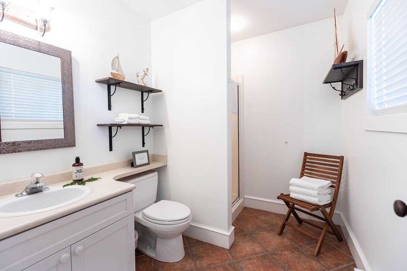 A bathroom with a sink, toilet, and a shower area. There are shelves above the sink and a chair with towels.