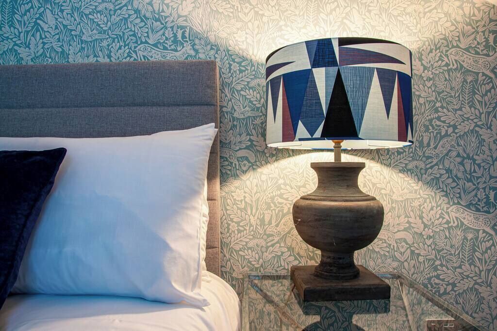 A bed with a white pillow and a dark blue pillow, a patterned wall, and a lamp with a geometric design on a glass table.