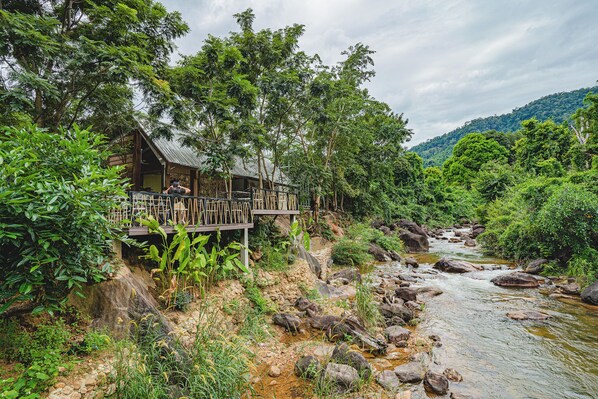 A wooden cabin with a deck overlooking a river, surrounded by lush greenery and a mountainous backdrop.