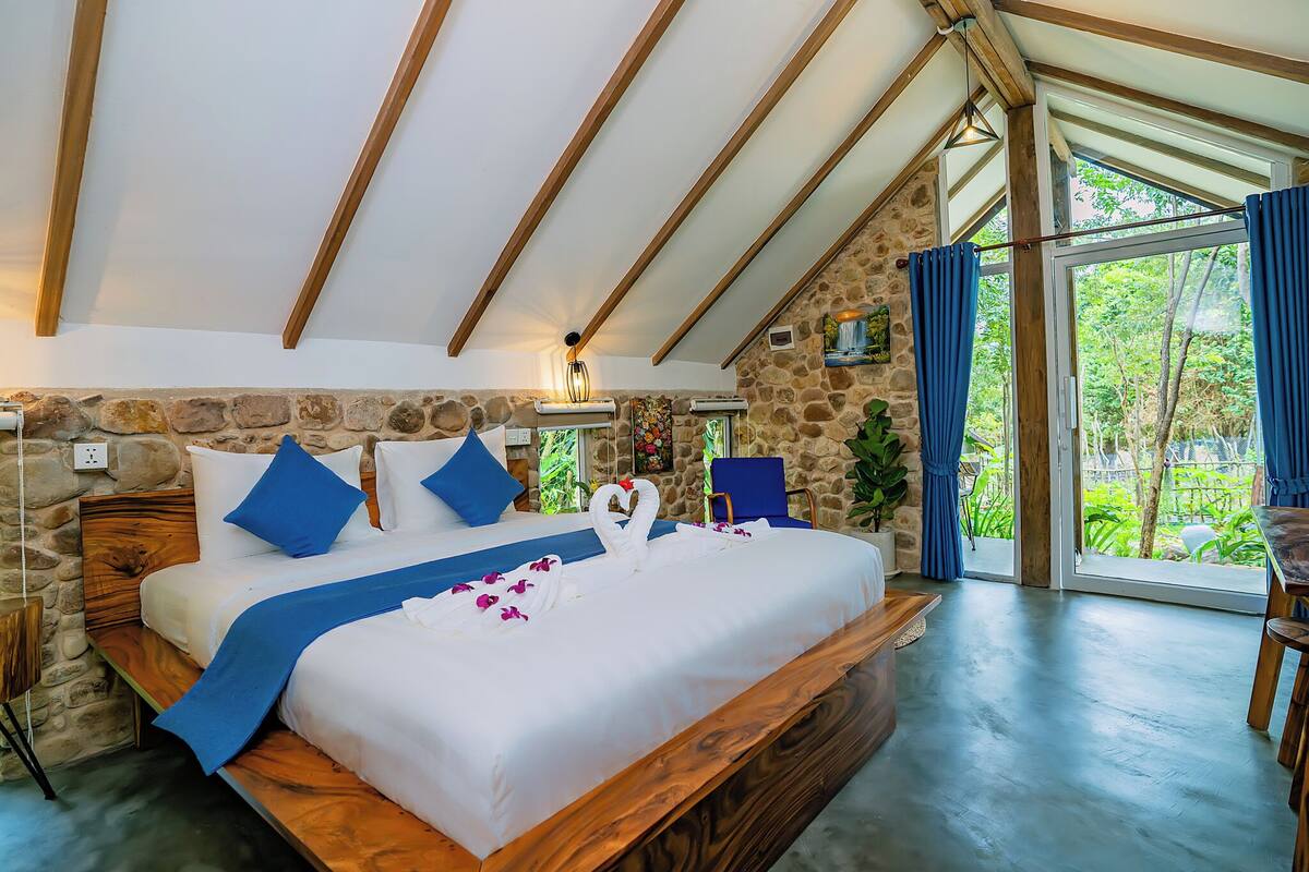 A bedroom with a stone wall, wooden ceiling beams, a large bed with blue pillows, and a view of greenery through a glass door.