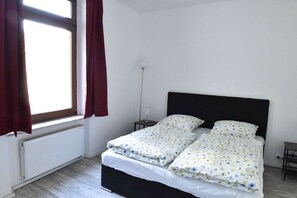 A small, single-bed room with a white radiator, a bedside table with a lamp, and a window with red curtains.
