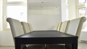 Un espace repas comprenant une table rectangulaire et six chaises, un mur blanc avec une partie recouverte de carreaux, et de grandes fenêtres laissant entrer la lumière naturelle.