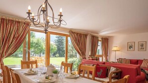 A dining area with a table set for four, wooden chairs, a chandelier, and large windows with curtains. - Chesa Dal Luf B1 by Interhome (Silvaplana-Surlej)
