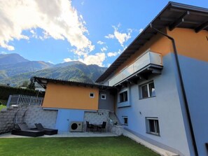 Una casa moderna de dos pisos con un muro de piedra y fachada azul y naranja, con montañas al fondo.