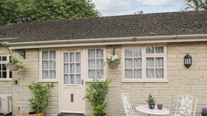 A stone cottage with a white door, two potted plants, a white chair, a small round table, and a hanging flower basket. - Paddock Cottage (Malmesbury)