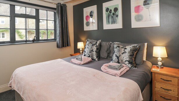 A bedroom with a bed, two bedside tables, a window with curtains, and three wall-mounted artworks. - Paddock Cottage (Malmesbury)
