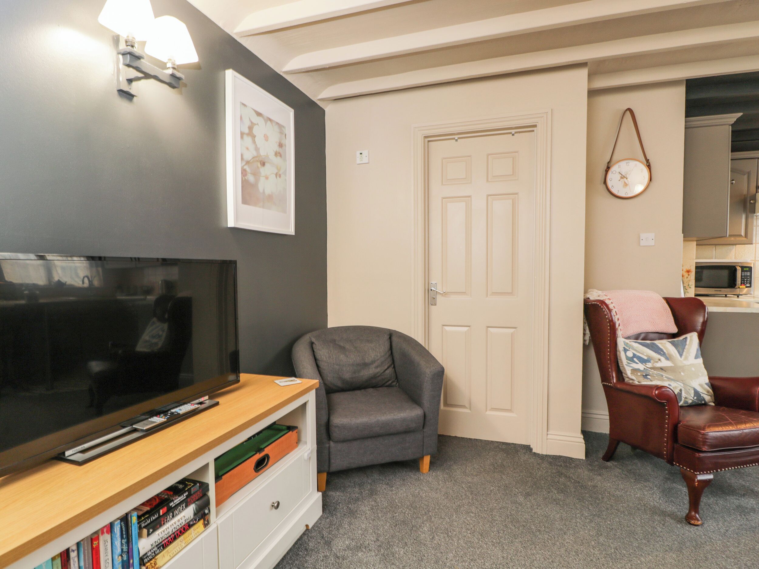 Una sala de estar con un televisor de pantalla plana, un sillón gris, un sillón de cuero marrón, una estantería y una puerta.