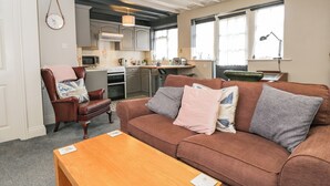 A cozy living room with a brown sofa, a wooden coffee table, and a dining area with a kitchen in the background. - Paddock Cottage (Malmesbury)