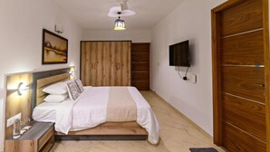 A bedroom with a bed, bedside table, lamp, ceiling fan, TV, wooden wardrobe, and a painting on the wall. - Lotus & Orchid Villas by Ramnath Homes (Alto de Porvorim)