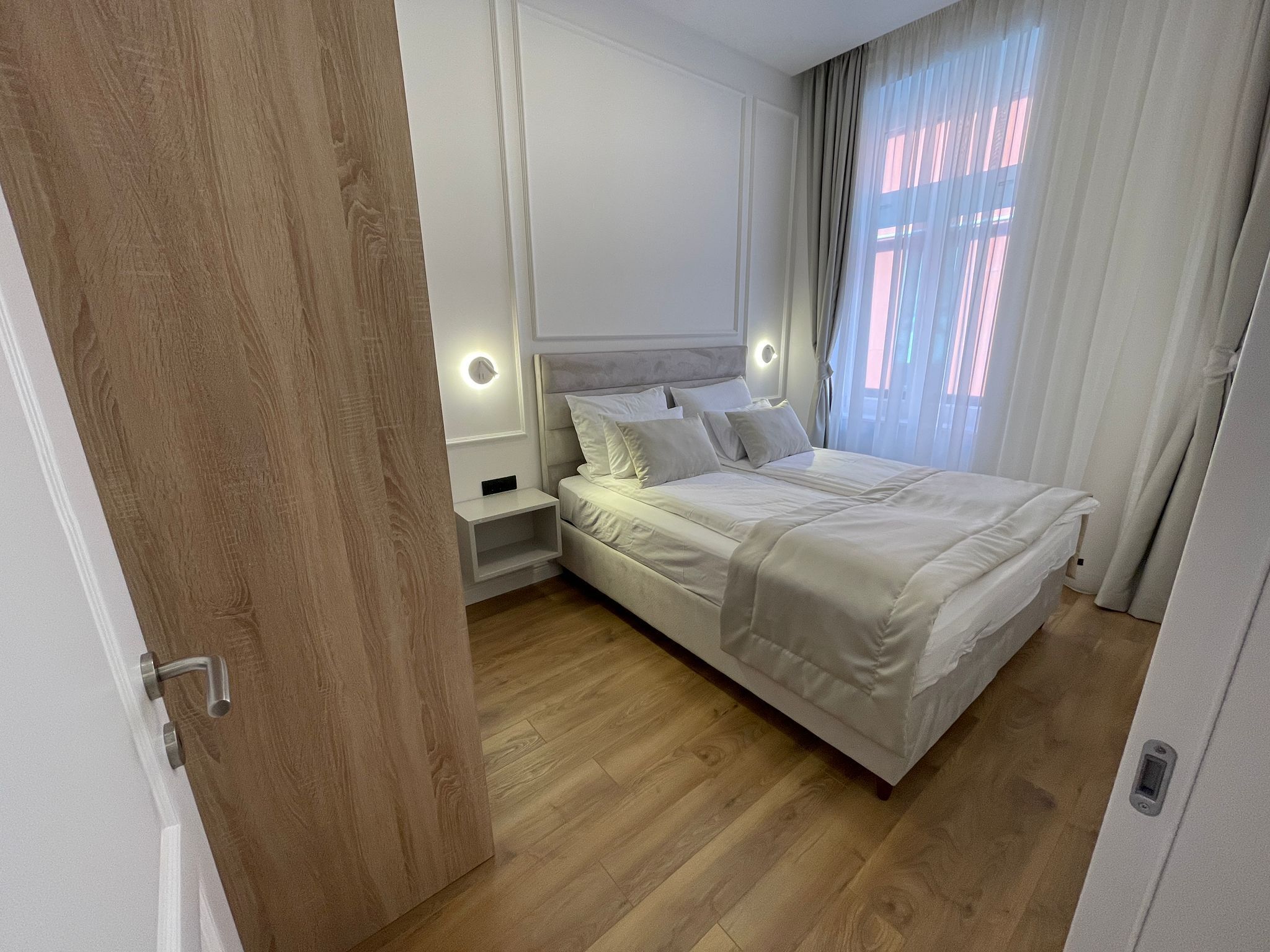 Una camera da letto moderna con pavimento in legno, un letto grande con lenzuola bianche, due comodini con luci a parete e una finestra con tende leggere.