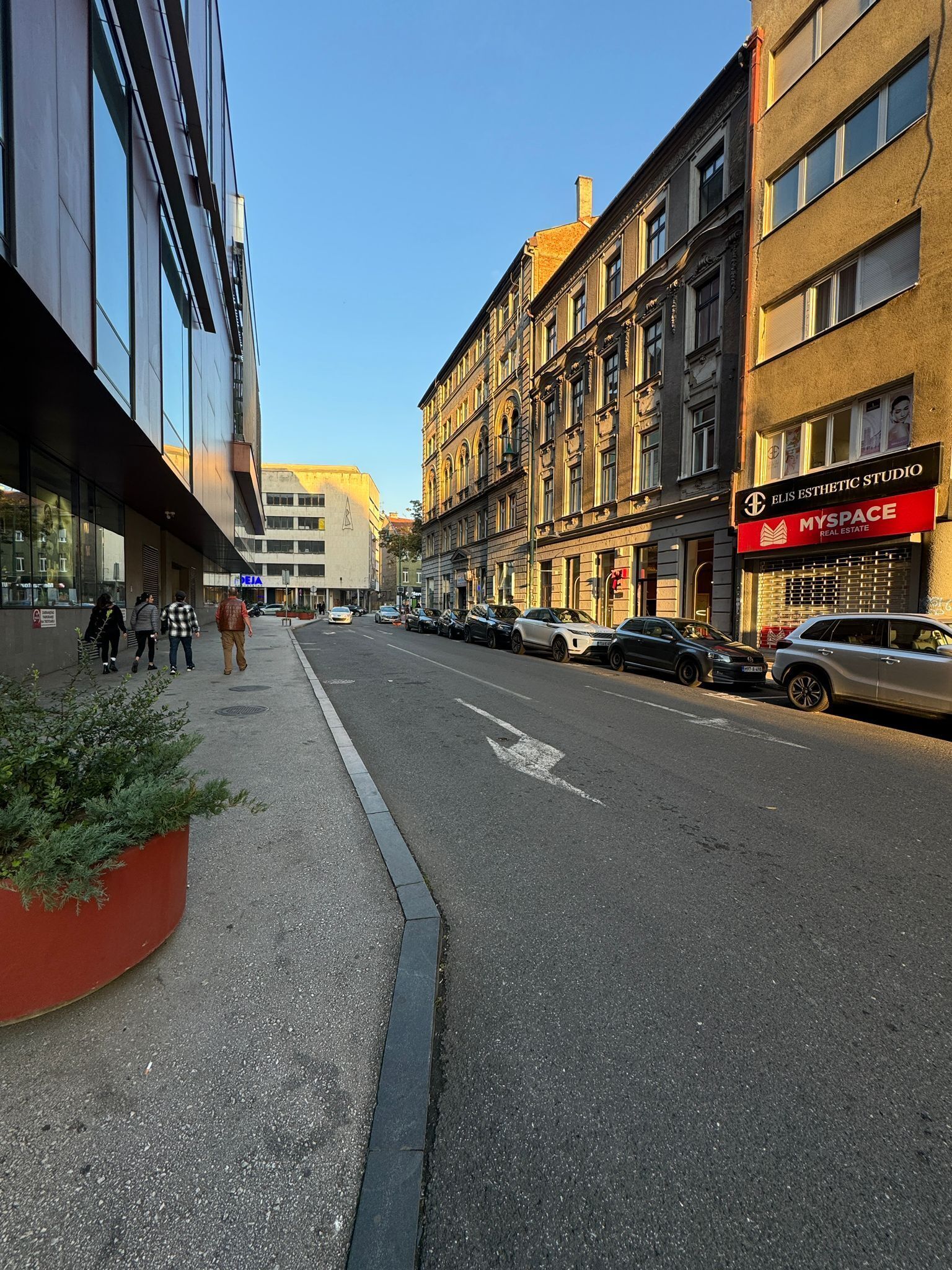 En gade med både moderne og historiske bygninger, klar himmel og parkerede biler.